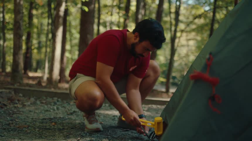 A bearded man hammers a metal stake into the ground to secure his tent at a campsite in the woods