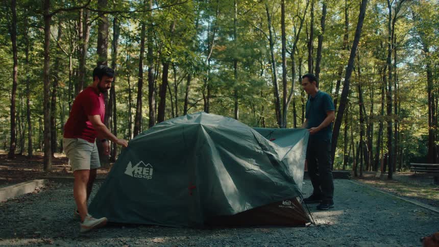 Two male friends work together in slow motion to put the rainfly on their tent at a sunny forest campsite
