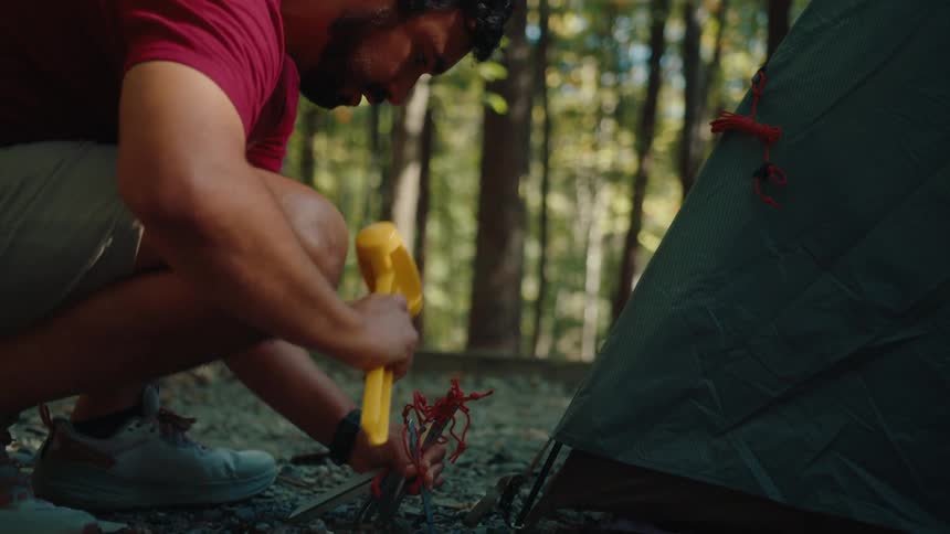 A male camper uses a yellow mallet to secure tent stakes into the ground at a campsite in the woods