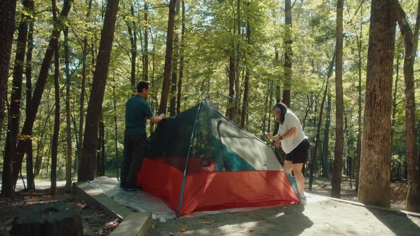 A man and woman work together to assemble their tent at a campsite in a beautiful sunlit forest