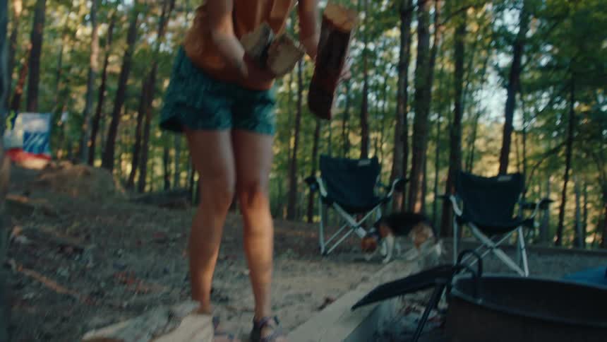 A woman in a cap gathers logs and carries them to a fire pit at a campsite in the woods in slow motion