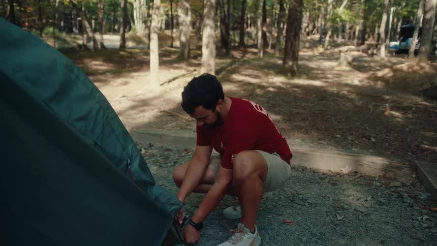 A young man carefully assembles his tent at a scenic campsite surrounded by trees