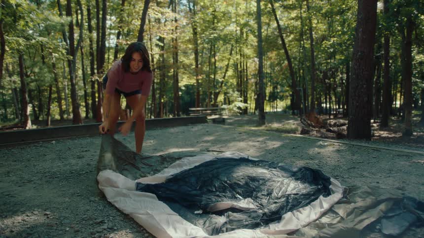 A young woman unfolds the fabric of her tent in slow motion while setting up camp in a sunny forest