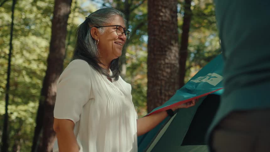 A smiling older woman happily helps her partner set up a tent while camping in a sunny forest