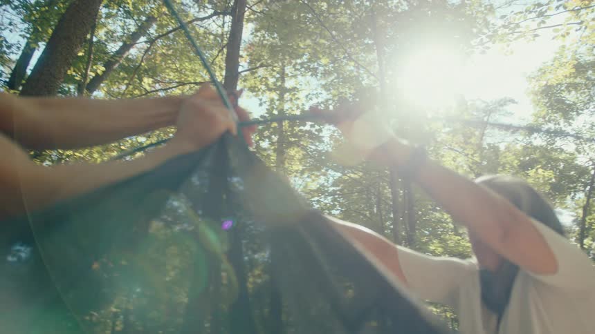 Slow motion, low-angle shot of a couple working together to assemble a tent at a sun-drenched campsite in the woods