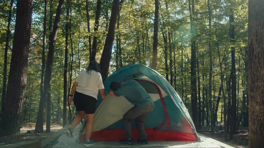 A couple sets up their campsite by assembling a tent together in a sunny forest