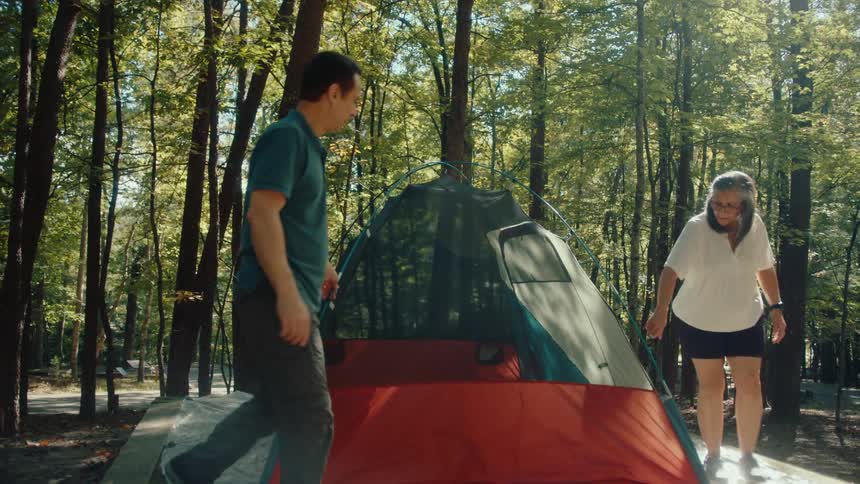 A middle-aged couple works together to set up their tent at a sunny campsite in the forest