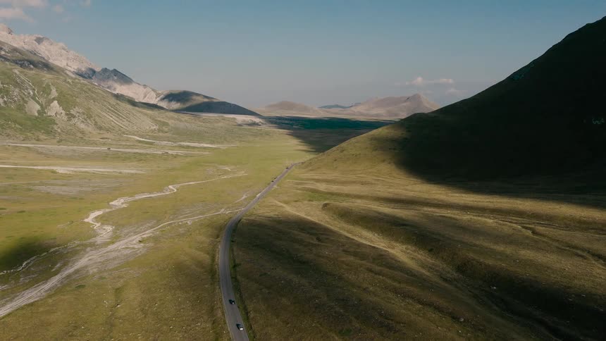 Cars travel on a remote road winding through a magnificent green valley nestled between mountains under a clear blue sky