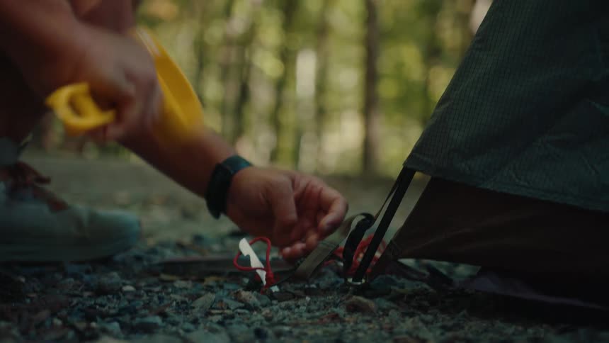 A tired hiker in slow motion hammers a tent stake into the rocky ground while setting up camp in the forest