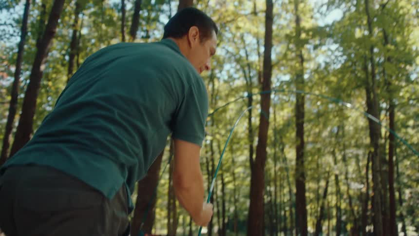A couple works together in slow motion to assemble their camping tent at a sunny campsite in the forest