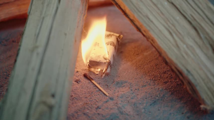 A slow-motion close-up of a fire starter burning in a sandy pit, preparing to ignite logs for a campfire