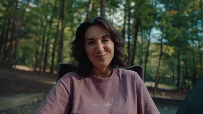 A happy young woman smiles warmly at the camera before turning to peacefully enjoy her campsite in the serene forest