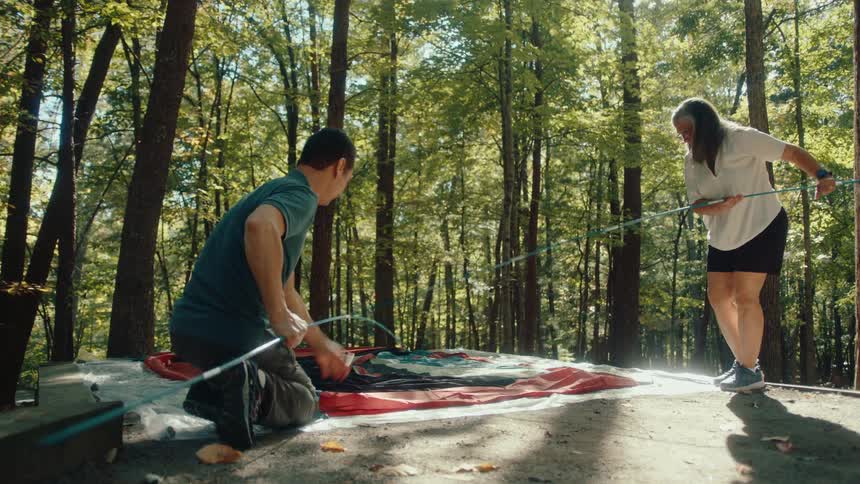 A couple collaborates on setting up their tent for a camping trip in a sunny forest