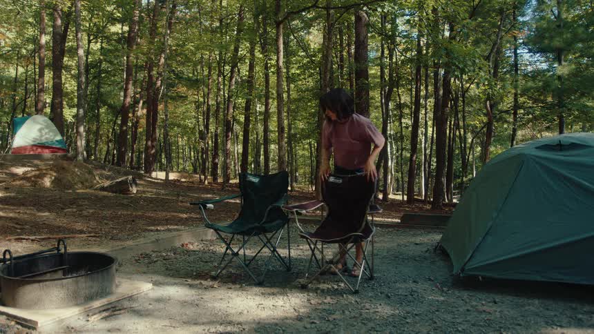 A young couple sets up their camp chairs and tent at a beautiful wooded campsite for a weekend getaway