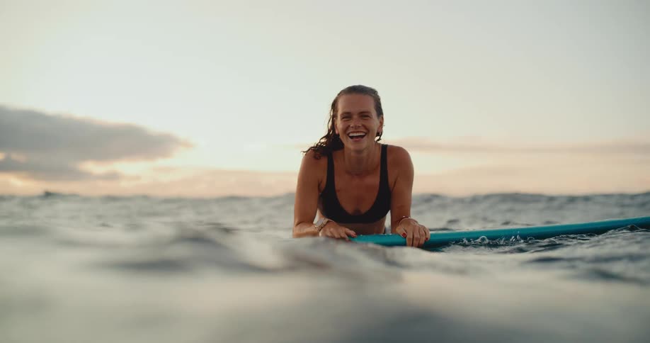 A smiling woman floats on her surfboard in the gentle ocean waves, laughing happily at sunset