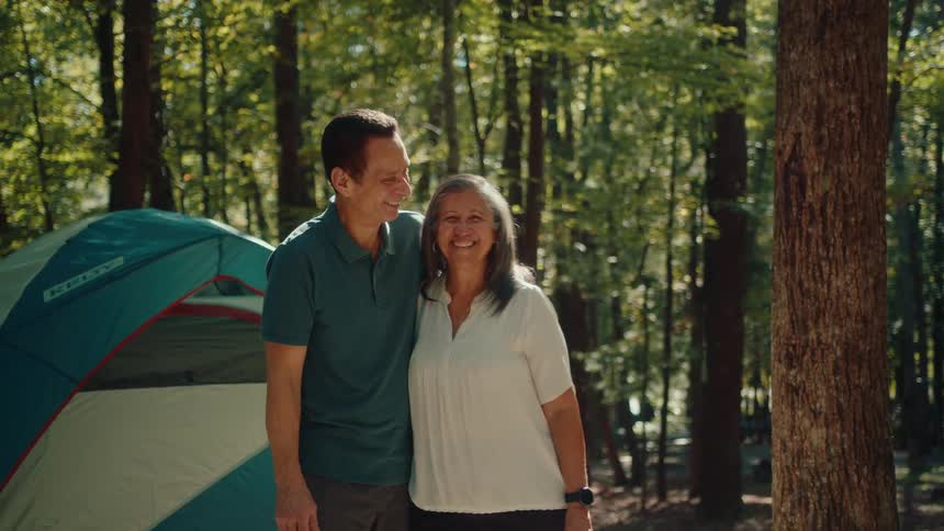 A happy senior couple smiles lovingly at each other while posing for a photo in front of their tent at a sunny forest campsite