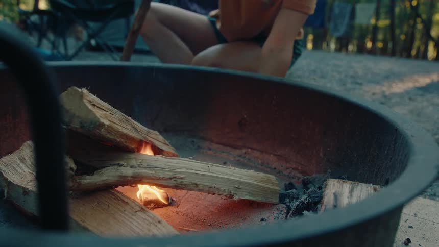 A woman in a cap carefully blows life into a campfire at a forested campsite in slow motion
