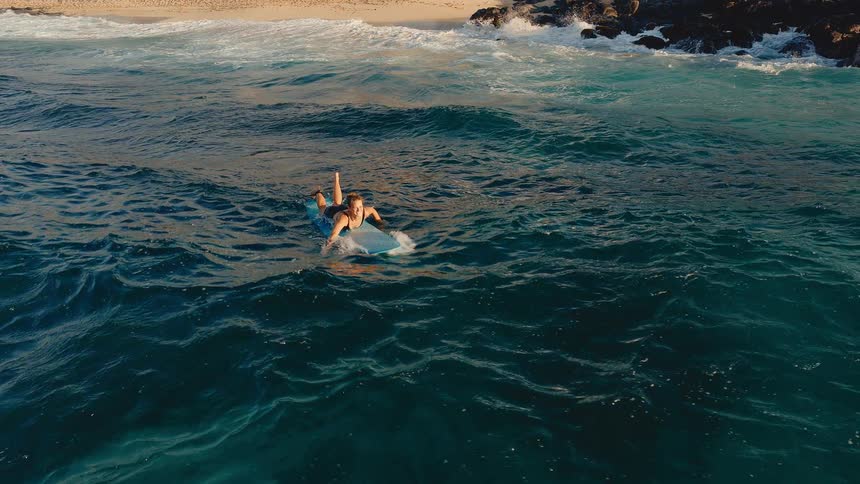High angle slow motion shot of a woman paddling out on her surfboard in the turquoise ocean water