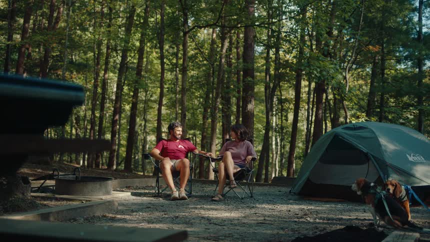 A joyful couple and their dogs arrive at a peaceful forest campsite, high-five in their chairs, and celebrate the start of their camping trip