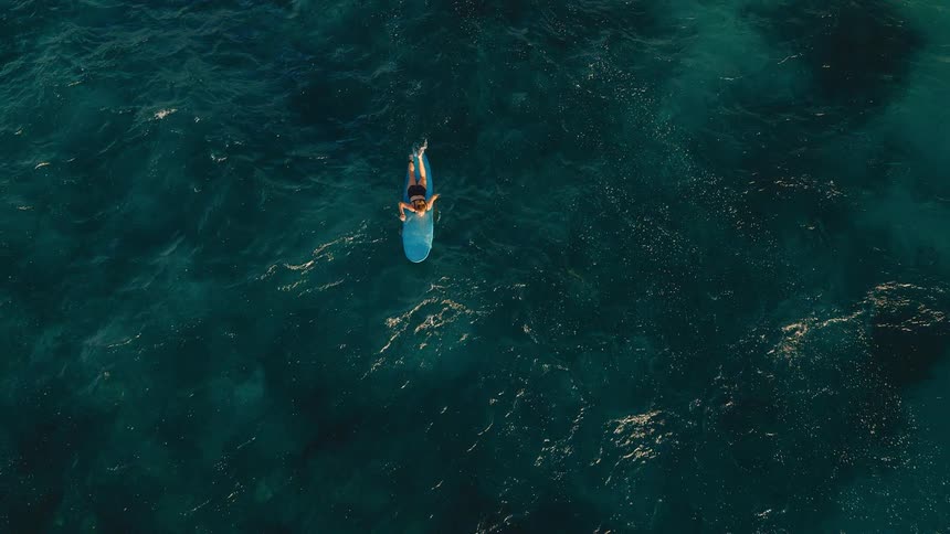 Aerial top-down shot of a lone surfer peacefully paddling on their board in the vast, deep turquoise ocean