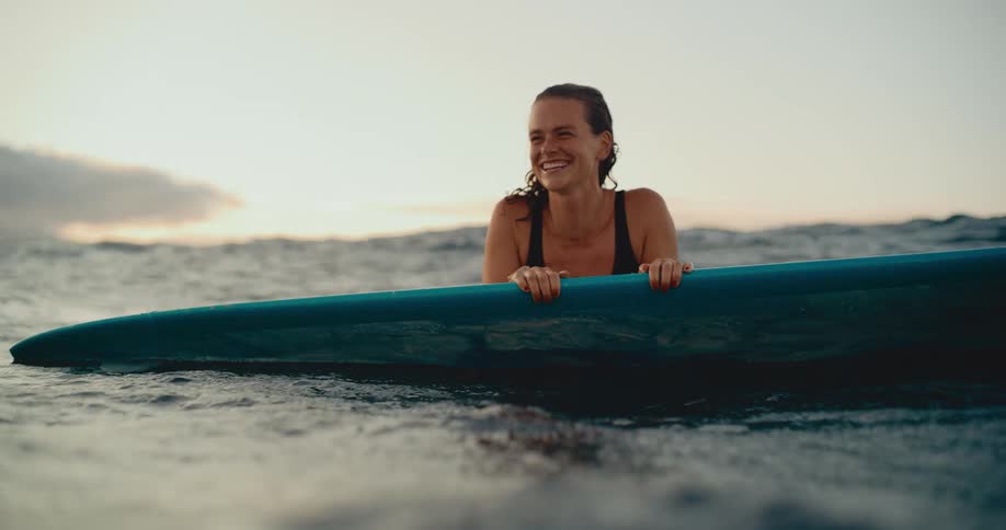 An attractive young woman with wet hair smiles contentedly while lying on her surfboard at sunset