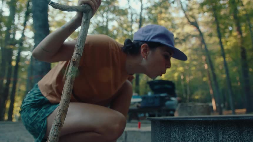 A woman in a baseball cap crouches and blows on a fire pit to start a campfire at a sunny wooded campsite in slow motion
