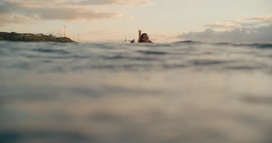 A slow motion, water-level view shows a smiling surfer woman paddling on her surfboard in the ocean at sunset