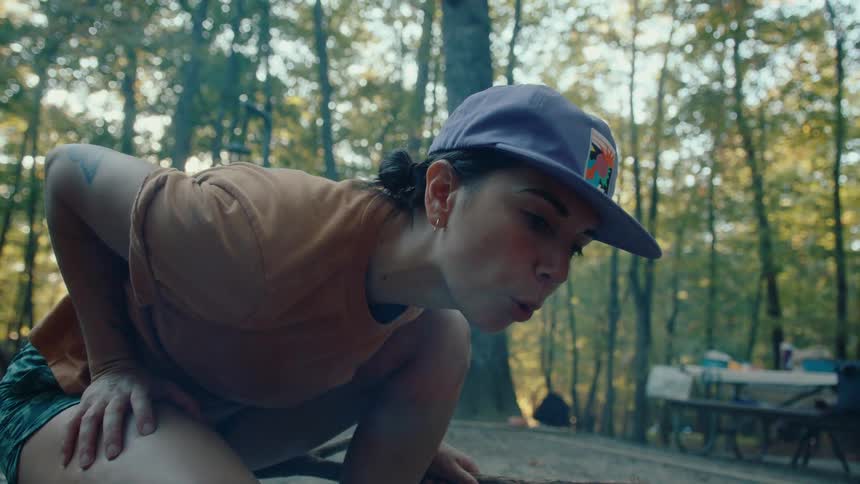 A woman in a baseball cap crouches down and gently blows on a small flame to start a campfire at a wooded campsite