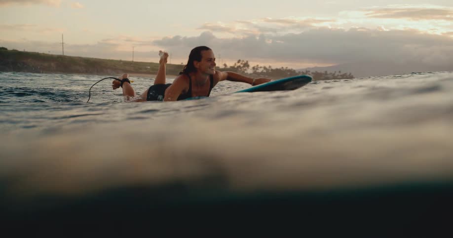 A smiling woman paddles her surfboard on the ocean at sunset, filmed in beautiful slow motion from the water's surface