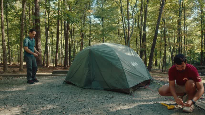 Two men work together to set up their tent at a forested campsite on a sunny day