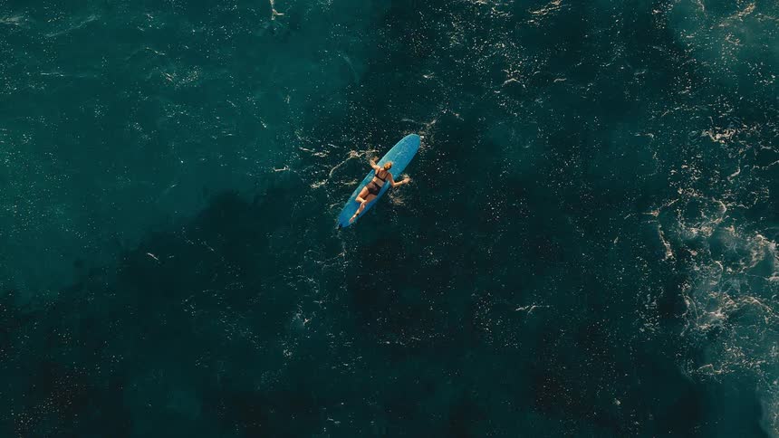 Aerial top-down shot of a woman in a bikini relaxing on her surfboard in the deep blue ocean
