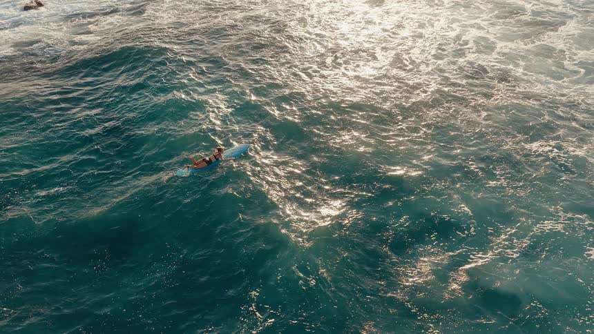 Static aerial top-down view of a surfer girl in a bikini paddling on a surfboard in the shimmering ocean
