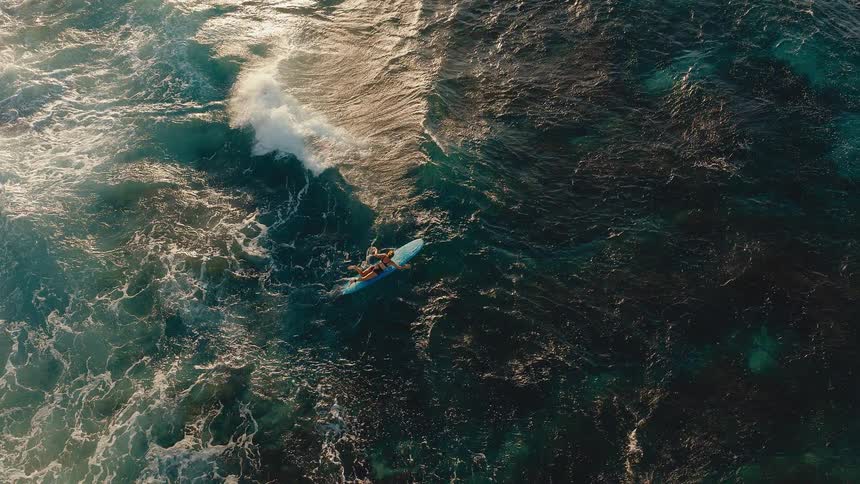 Aerial cinemagraph of a surfer paddling on a surfboard in a turbulent ocean at sunrise