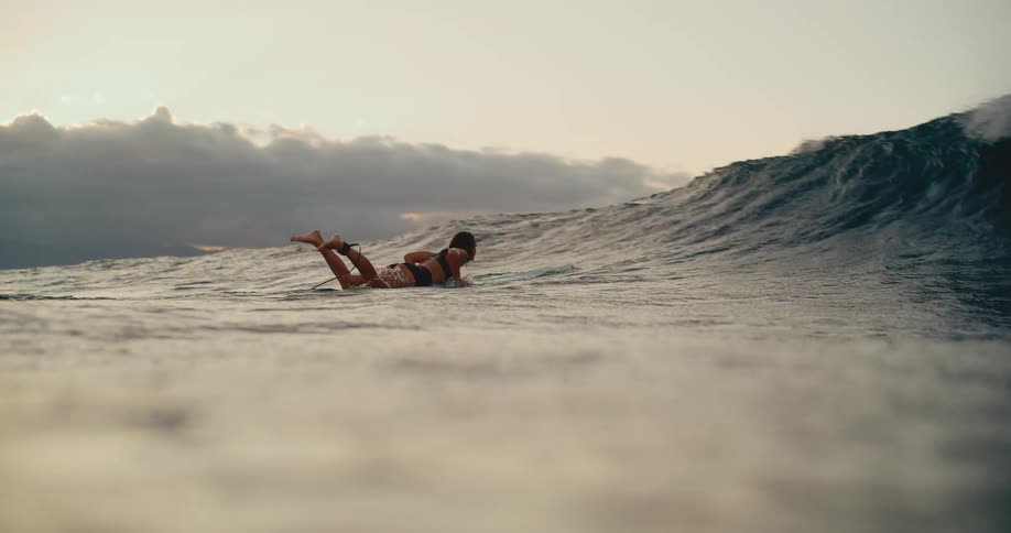 A female surfer paddles on her surfboard in slow motion as a powerful ocean wave breaks over her at sunset
