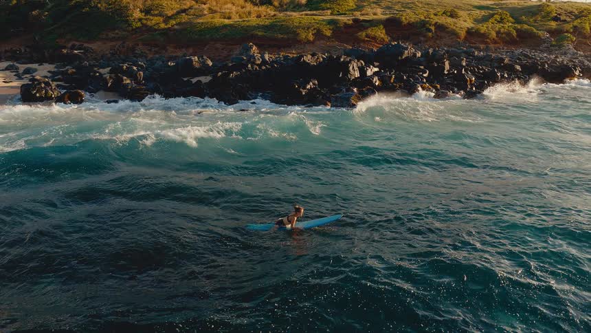 Aerial view of a female surfer in a bikini paddling out on her longboard in the deep blue ocean near a rocky coastline