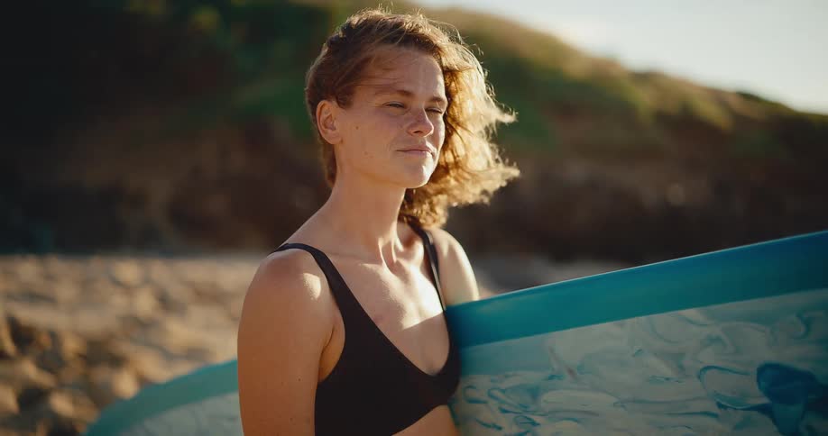 Portrait of a beautiful young woman surfer standing on the beach holding her surfboard at sunset, looking out at the ocean with the wind in her hair