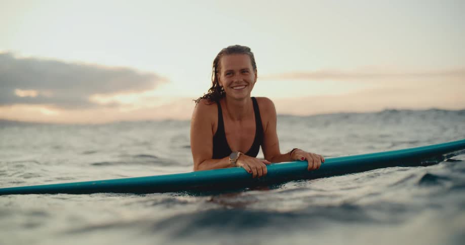 Slow-motion portrait of a smiling woman lying on her surfboard in the ocean at dusk