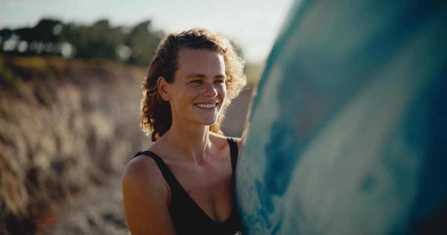 A radiant young woman with freckles smiles warmly while holding her surfboard, bathed in the golden light of sunset on the coast