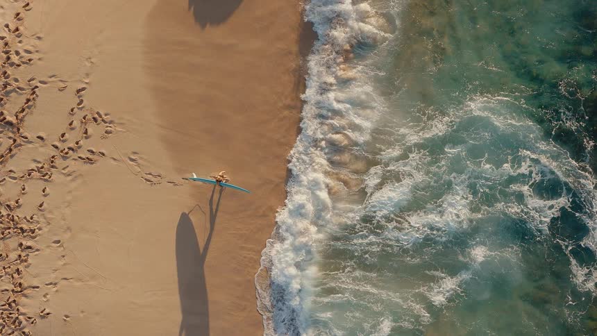 Top-down aerial slow-motion view of a surfer lying on their board at the edge of a sandy beach as waves wash ashore