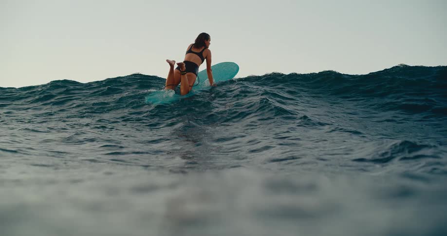 A female surfer in a bikini paddles over a large ocean wave as the camera dips underwater in slow motion