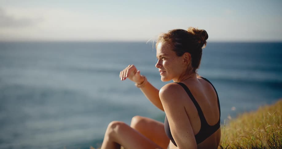 A beautiful young woman in a bikini sits on a grassy cliff and peacefully watches the ocean waves in slow motion