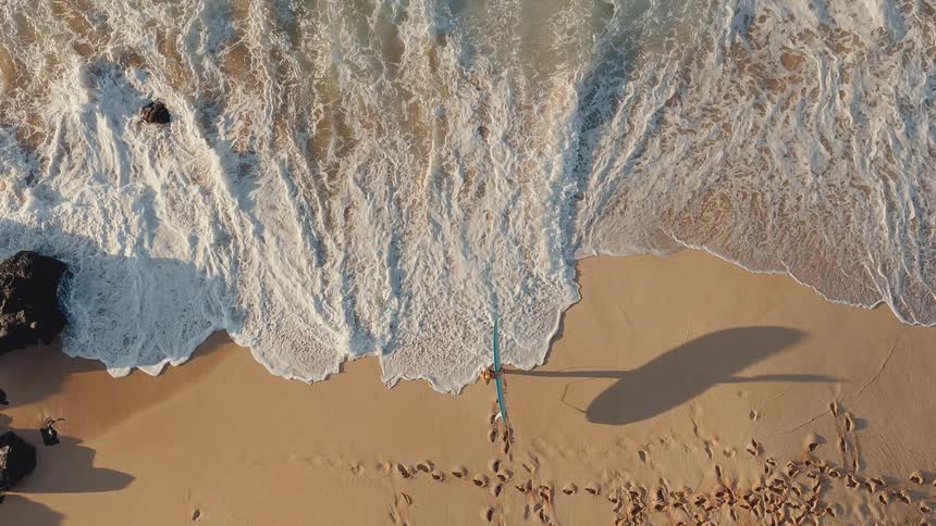 Slow motion top-down aerial view of a lone surfer standing on a golden beach watching the waves crash
