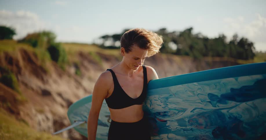A happy woman with short hair smiles as she walks with her surfboard, her hair blowing in the wind in beautiful slow motion