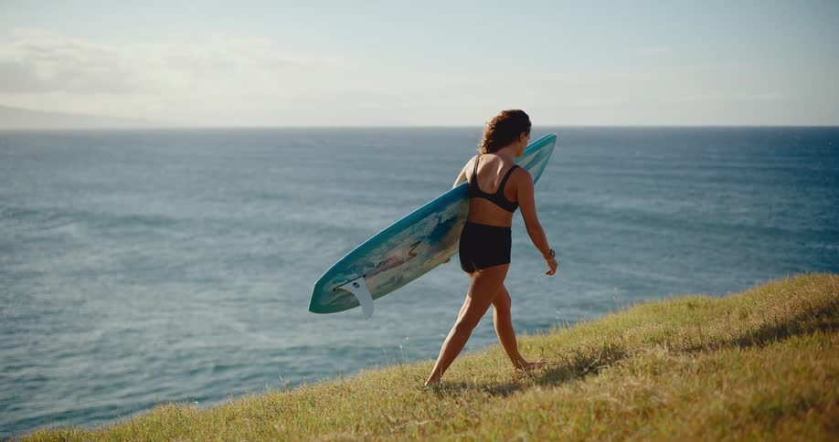 A female surfer walks up a grassy hill carrying her longboard while overlooking the vast ocean