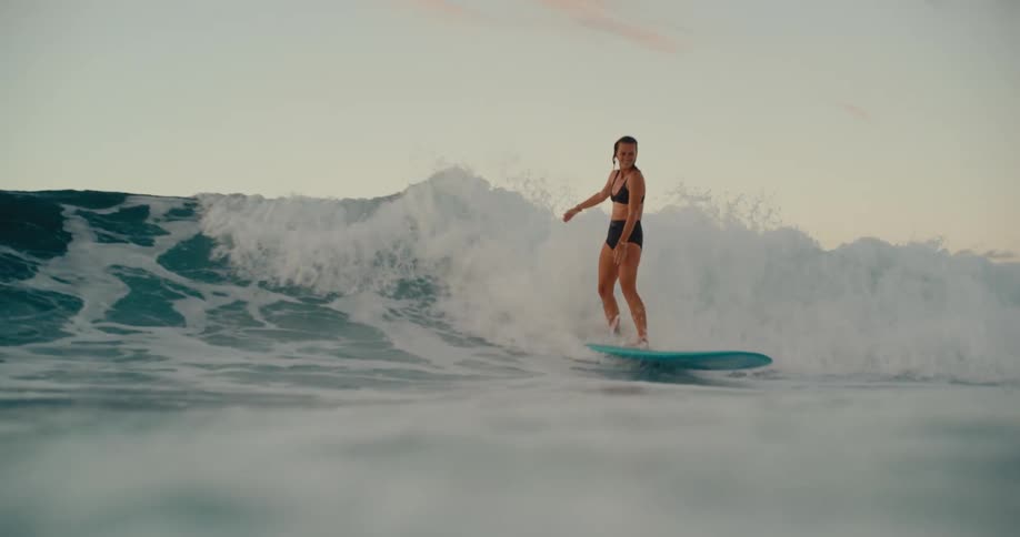 Young woman gracefully rides an ocean wave on her longboard at sunset, filmed in slow motion from a water-level perspective