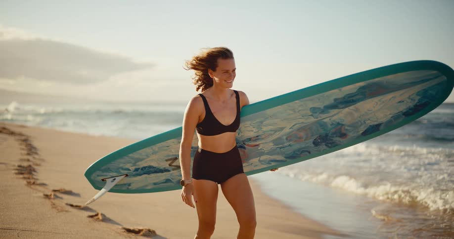 Happy female surfer walks down the beach with her surfboard at sunset in slow motion