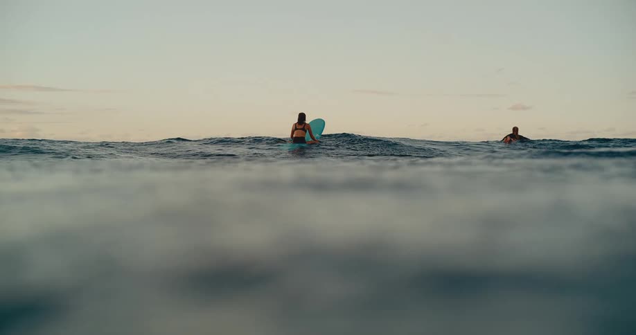 Two female surfers wait for waves on their surfboards in the ocean at dusk before the camera dips below the surface