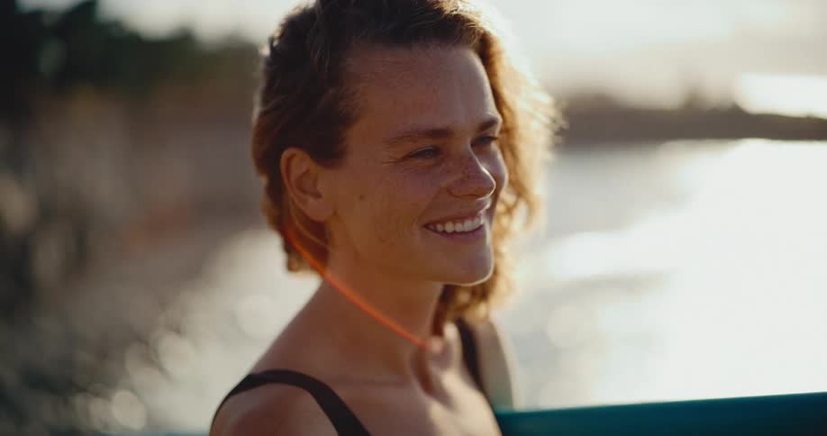 A close-up portrait of a happy female surfer smiling as she watches the ocean waves during a golden hour sunset