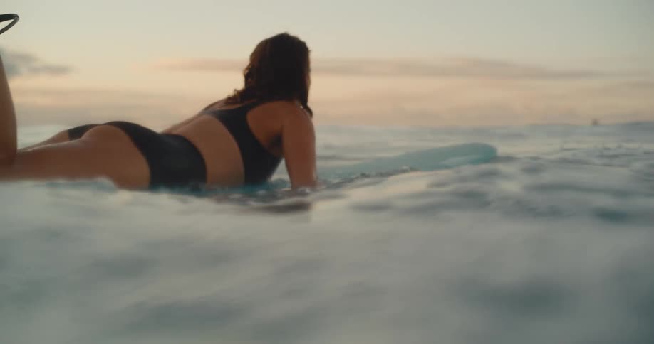 An athletic woman paddles on her surfboard in the calm ocean during a beautiful golden sunset, filmed in slow motion