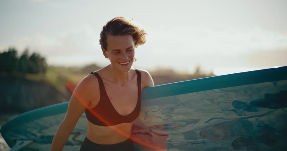 Slow motion close-up of a happy female surfer walking with her board along the beach at sunset